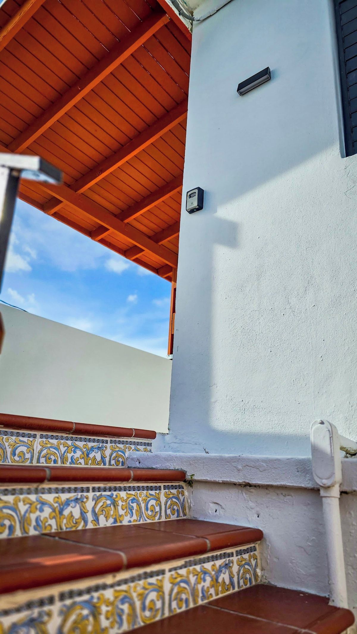 Un espacio al aire libre techado, con techo de madera, pared blanca con dispositivo de seguridad y piso de baldosas con diseño decorativo.