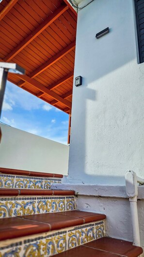 A covered outdoor area with a wooden ceiling, a white wall with a security device, and a tiled floor with a decorative pattern.