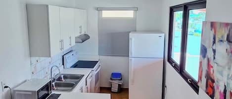 A compact kitchen with a sink, stove, and refrigerator, a small dining area with a chair, and a window with a view.