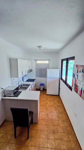 Cocina compacta con lavamanos, estufa y refrigerador, un pequeño comedor con una silla y una ventana con vistas.