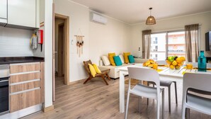 A modern kitchen with white cabinets, a wooden countertop, and a built-in oven. There is a dining area with a white table, chairs, and a fruit bowl. A sofa with colorful cushions is visible in the background. - Bright Apartment with Pool in Portimão | 5 Min Walk to the Beach by Lisbeyond (Portimão)
