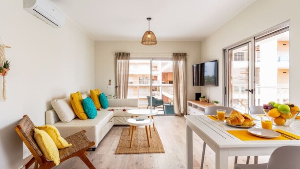 A modern living room with a sofa, a dining table with breakfast items, a TV, and a balcony with curtains. - Bright Apartment with Pool in Portimão | 5 Min Walk to the Beach by Lisbeyond (Portimão)