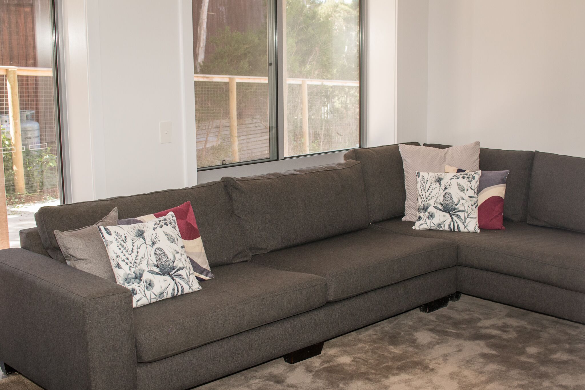 A grey sectional sofa with floral and geometric patterned cushions, positioned in a room with large windows and a view of a wooden deck.
