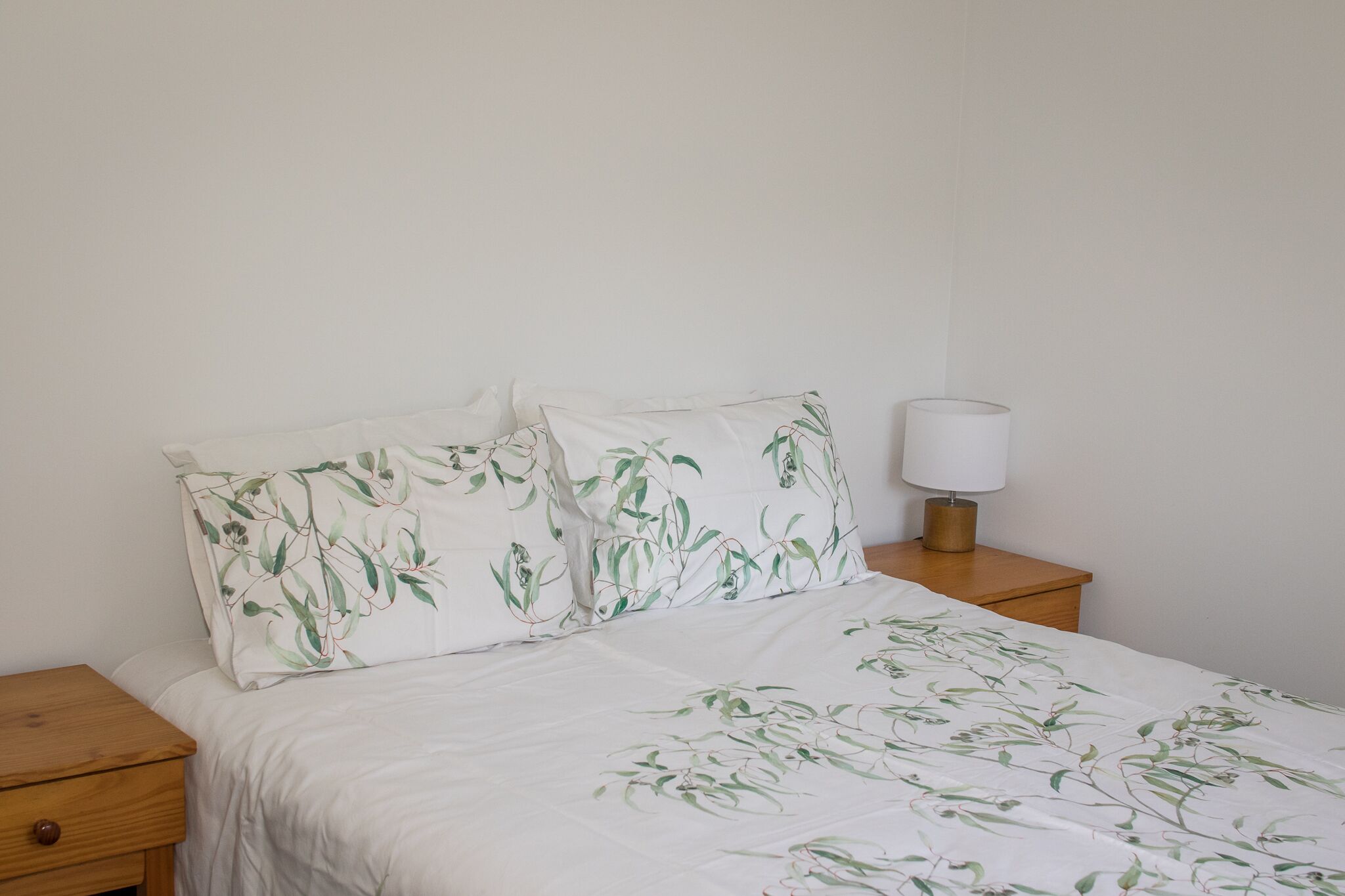 Un lit recouvert d’une couvre-lit à motifs floraux, deux oreillers au même motif, une table de chevet avec une lampe et une petite table en bois placée à côté du lit.