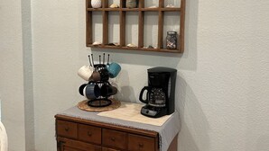 A kitchenette with a coffee maker, a shelf with decorative items, and a wooden cabinet. - Beachy OBX 2Bedrm-No Stairs-Close to beach and shops! (Kill Devil Hills)
