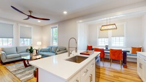 Una cocina moderna con una isla central, un comedor con sillas naranjas y una sala de estar con un sofá gris.