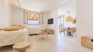 A modern bedroom with a bed, bedside table, and a window. There is a dining area with a table and chairs visible in the background. - Casa del Mare Blu - Seaview apartment with pool (Tertenia)