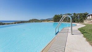 A clear blue swimming pool with a metal ladder, surrounded by a concrete deck and a grassy area with trees and hills in the background. - Casa del Mare Blu - Seaview apartment with pool (Tertenia)