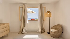 A bedroom with a bed, a wooden dresser, a wicker chair, a floor lamp, and a view of the sea through a window with curtains. - C'est la vue - Seaview apartment with pool (Marina, Tertenia)