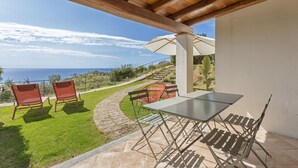 A covered patio with a table and chairs, overlooking a grassy area and the sea. - C'est la vue - Seaview apartment with pool (Marina, Tertenia)