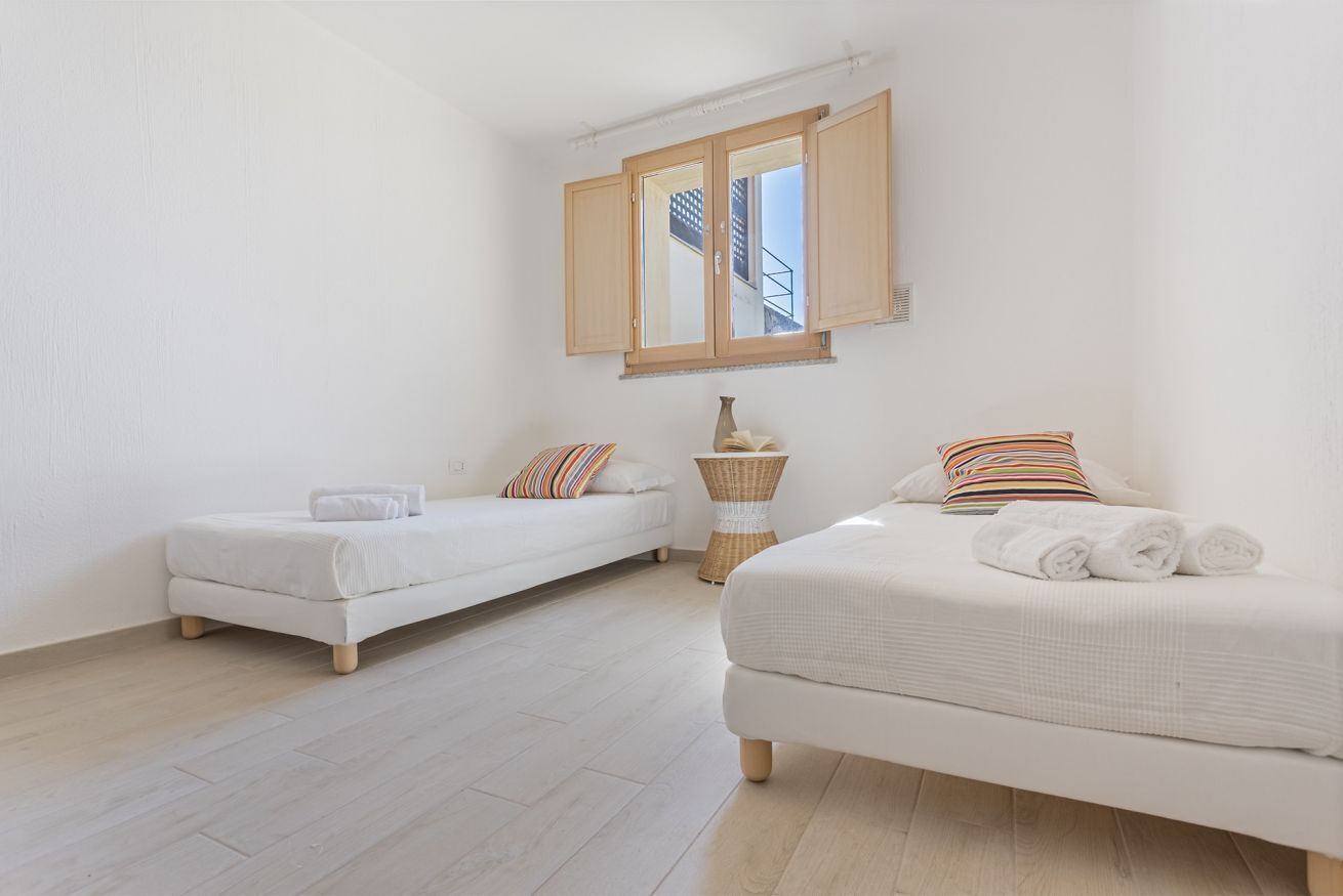 Une chambre avec deux lits simples, une grande fenêtre donnant sur un immeuble et une petite table d’appoint avec un vase.
