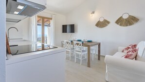 Moderni keittiö, jossa on sisäänrakennettu liesi, ruokailutila puupöydällä ja valkoisilla tuoleilla sekä valkoinen sohva, jossa on punaiset koristetyynyt.