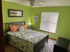 Une chambre à coucher comprenant un lit, une table de chevet, un ventilateur de plafond, une fenêtre avec des stores et un tableau encadré accroché au mur.
