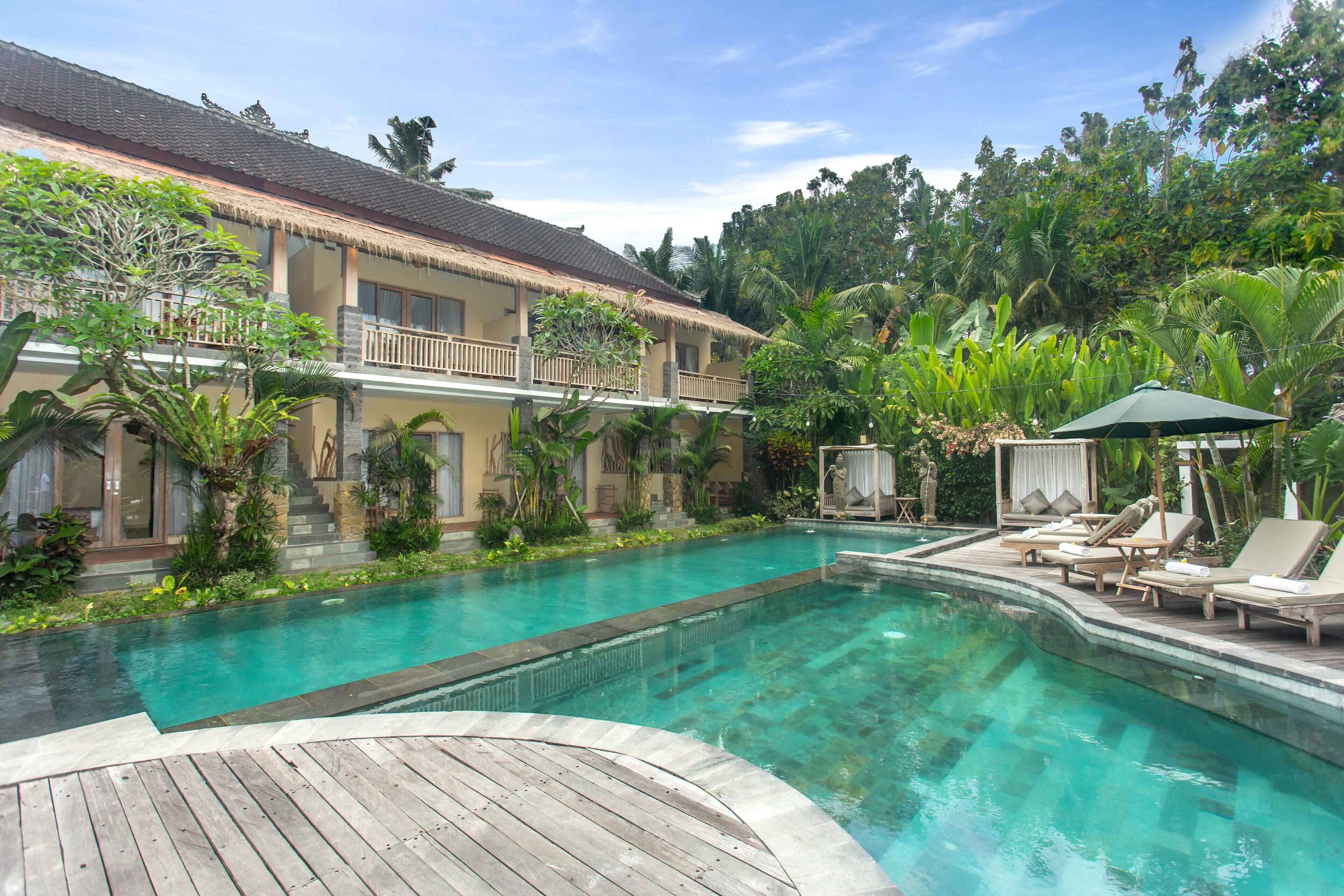 A pool area with a wooden deck, lounge chairs, and a building with a tiled roof and balconies.