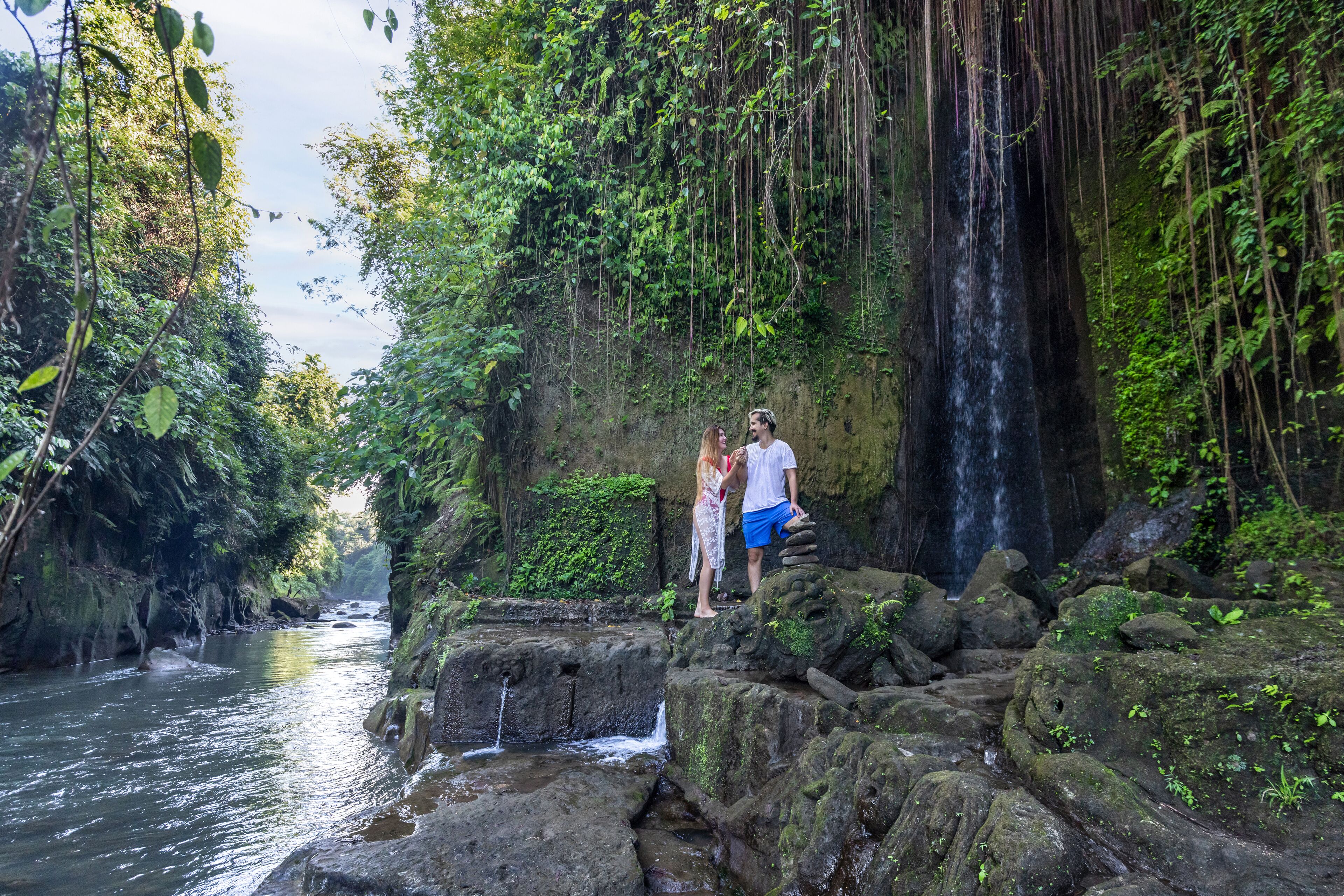 A couple standing on a rocky ledge overlooking a waterfall in a lush, green forest setting.