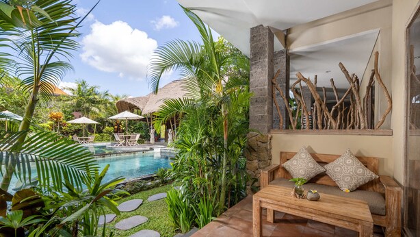 A poolside area with a wooden bench, lounge chairs, and a stone wall with decorative branches. - Kailash Suites Ubud (Sukawati)