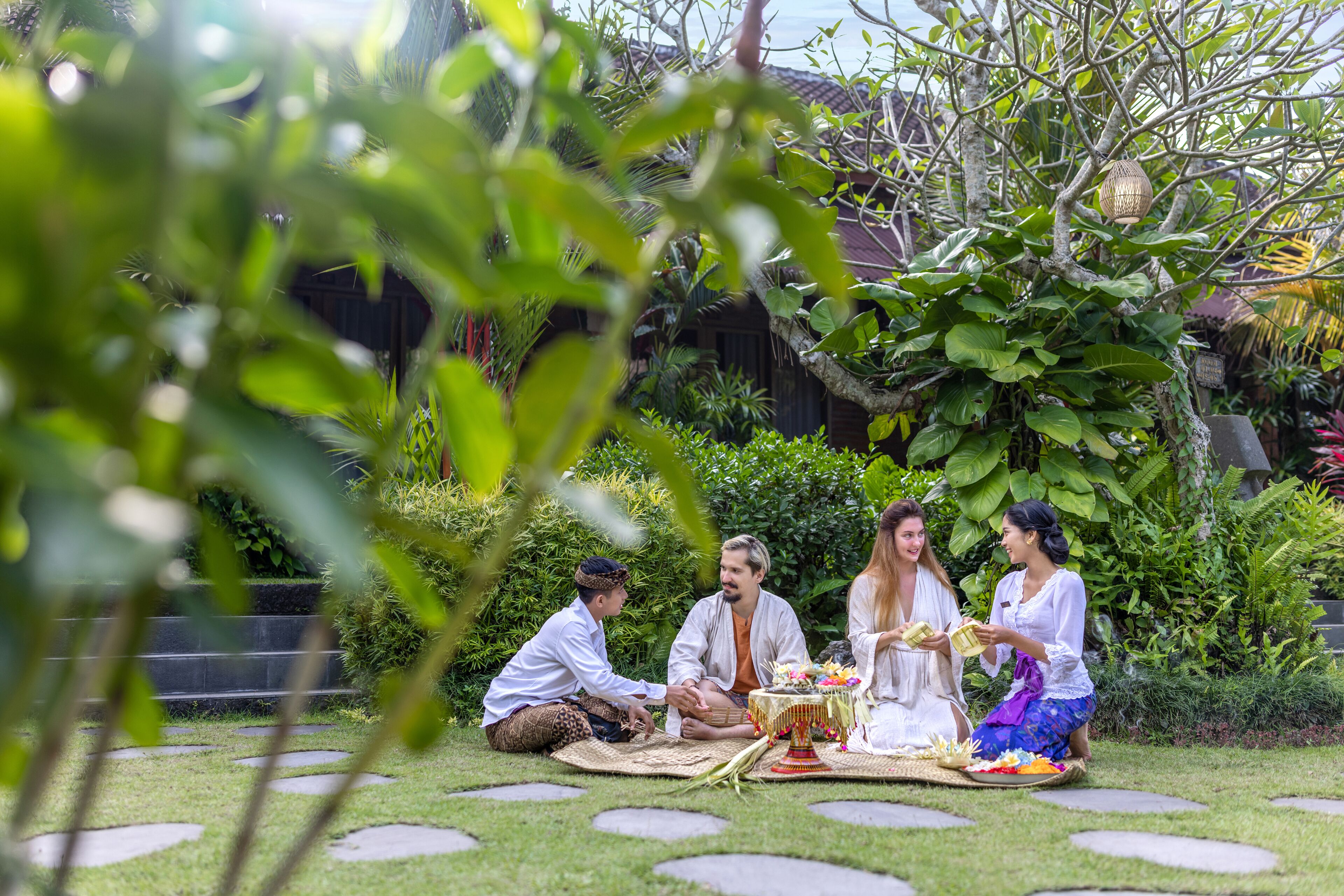A group of people seated on a mat outdoors, engaged in a meal with a table set with food and drinks.