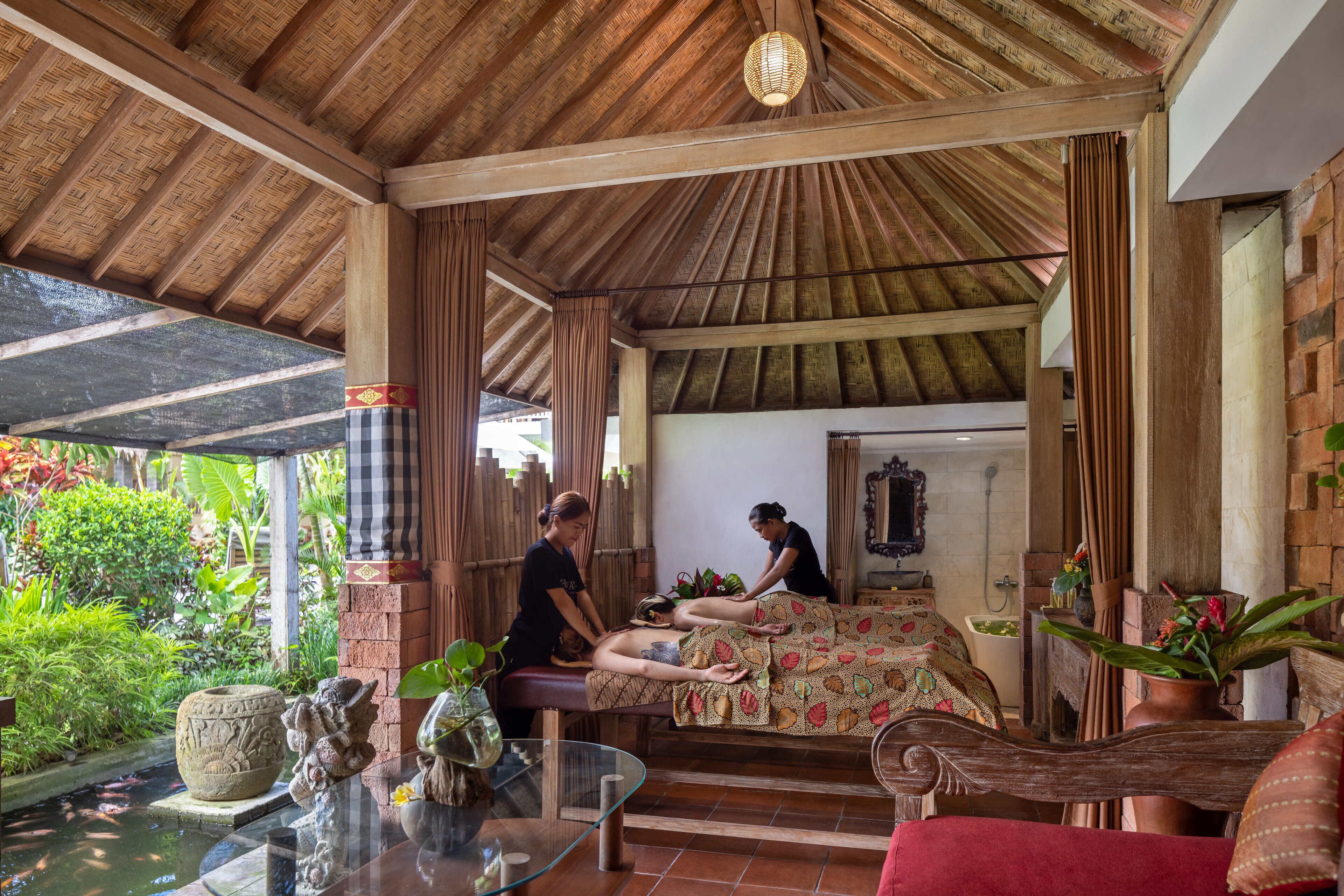A traditional spa setting with a wooden ceiling, a massage table, and a small garden area visible through the open sides.