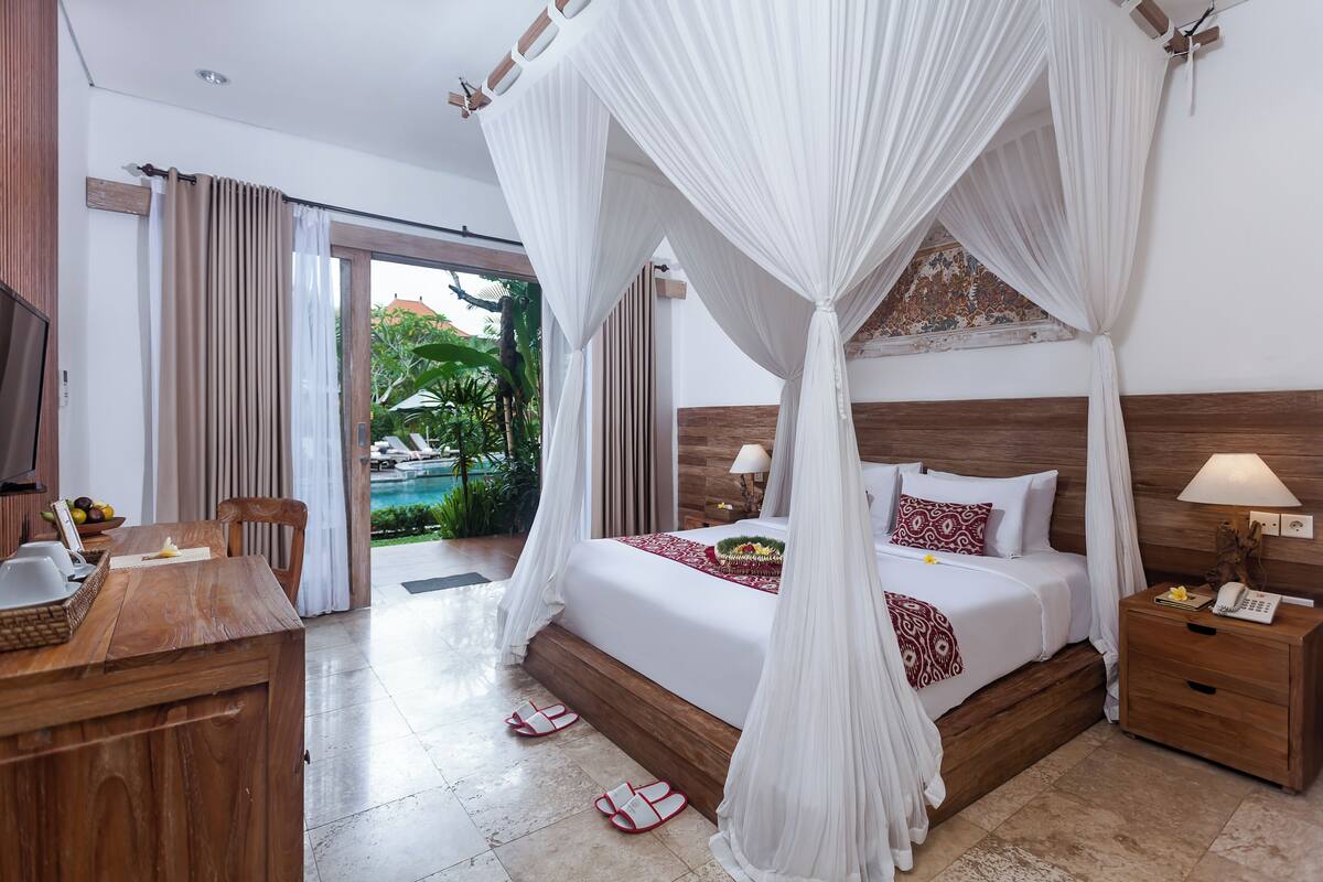 Habitación de hotel con cama con dosel, escritorio de madera, mesita de noche y vista a la piscina y follaje exterior.