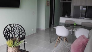 Una sala moderna con un sofá, una mesa de comedor con sillas, un televisor montado en la pared y una cocina visible a través de una puerta abierta.