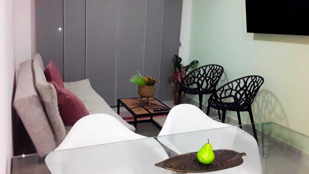 Una sala de estar moderna con un sofá, dos sillones blancos, una mesita con una planta y un televisor montado en la pared.
