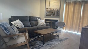 Uma sala de estar com sofá, mesa de centro de madeira, cadeira de balanço e uma cadeira. Há uma lareira com um quadro emoldurado acima.