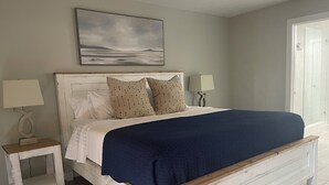 Quarto com cama, duas mesas de cabeceira, um criado-mudo, um quadro na parede e uma porta à direita.