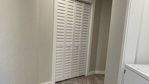 Um corredor com piso de madeira cinzento, paredes brancas e um armário branco com portas venezianas.