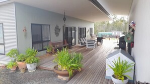 Ein überdachter Außenbereich mit Holzdeck, Kübelpflanzen, Grill und einer Sitzecke mit Stühlen und Tisch.
