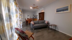 Ein Wohnzimmer mit grauem Sofa, einem kleinen runden Tisch und einem Holzstuhl. Es gibt einen Deckenventilator und ein Bild an der Wand.