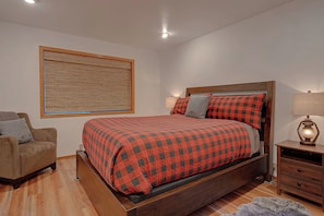 A bedroom with a bed, a chair, a bedside table with a lamp, and a large window with a blind. - Rogue Keystone Lodge/Epic River Views/Sleeps 7/SPA (Gold Beach)