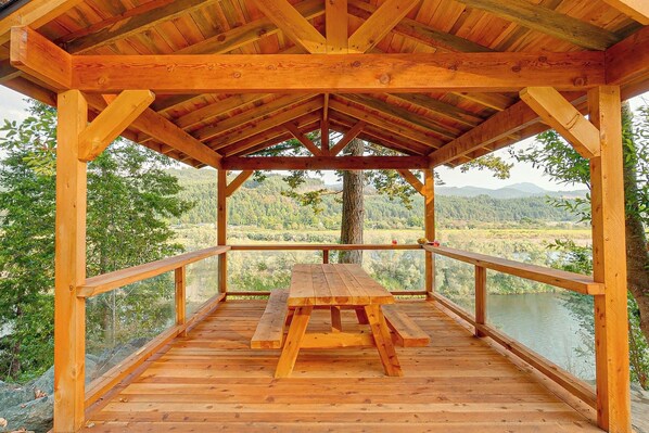 A wooden deck with a picnic table, surrounded by a railing, overlooking a lake and forested area.