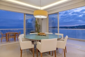 Uma sala de jantar moderna com mesa de vidro e cadeiras brancas, uma luminária pendente amarela e vista para o mar.