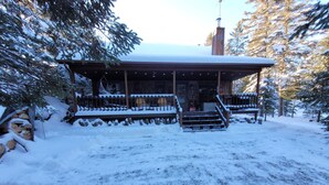 Una cabaña de madera con un porche cubierto y escalones que conducen a la entrada, rodeada de terreno y árboles cubiertos de nieve.