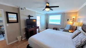 A hotel room with a large bed, a ceiling fan, a TV on a wooden dresser, a chair, a lamp, and a window with blinds. - Castaways 2c 2 Bedroom Condo by RedAwning (Gulf Shores)