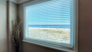 A window with horizontal blinds, a view of a beach, and a vase with dried plants on the windowsill. - Castaways 2c 2 Bedroom Condo by RedAwning (Gulf Shores)