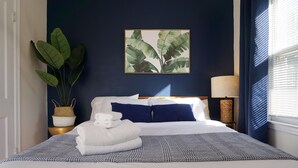 A neatly made bed with a blue and white patterned bedspread, a white pillow, and a blue pillow. There is a large potted plant to the left of the bed, a wall-mounted picture of leaves, and a lamp on a bedside table.