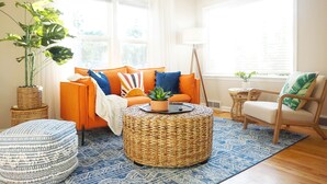 A living room with a sofa, ottoman, and chair, a round wicker coffee table, and a large window with curtains.