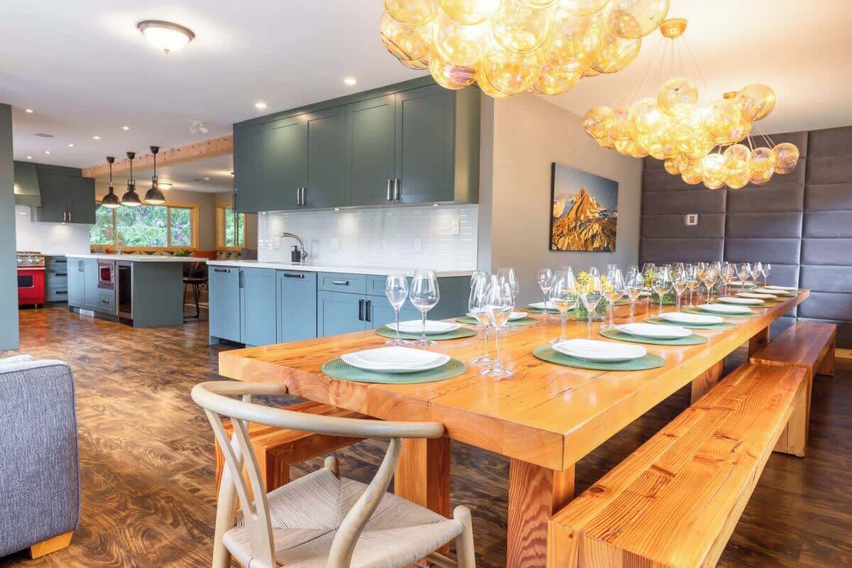 Cocina moderna con una larga mesa de comedor de madera dispuesta para una comida, con una lámpara colgante única y vistas al exterior.