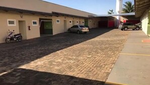 A parking area with a paved walkway, a scooter, and a car parked in front of a building with a flat roof and a water tank in the background. - hotel e motel ape goiania anel viario (Goiânia)