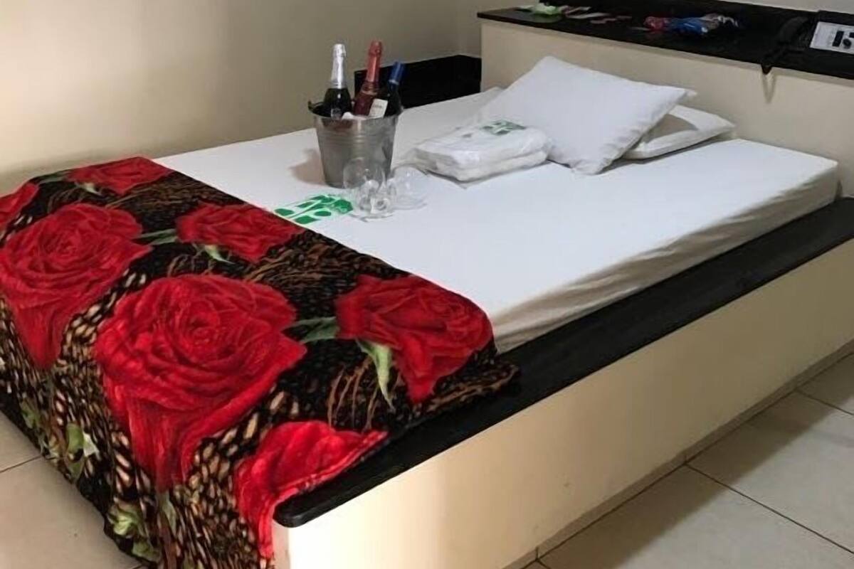 Ein Bett mit rotem Blumendecken, einem weißen Kopfkissen und einem kleinen Eimer mit Flaschen.