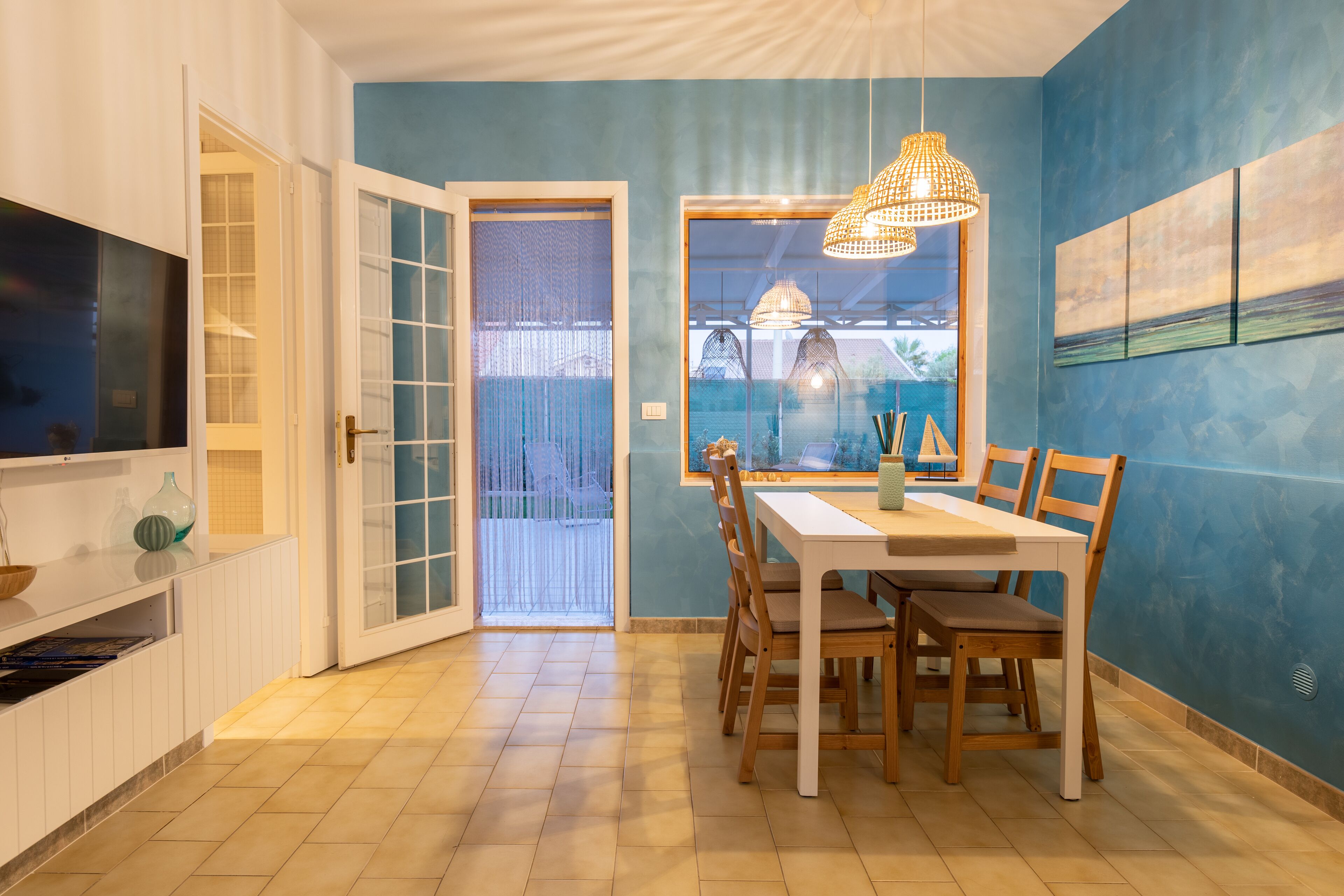 A dining area with a white table and chairs, a hanging light fixture, and a view through a window.