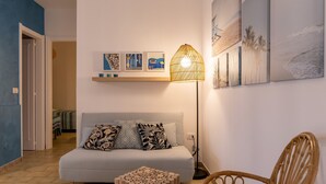 Ein modernes Wohnzimmer mit einer Couch, einem kleinen Holzschemel, einem Korbstuhl und einer Stehlampe. An den Wänden befinden sich Kunstwerke und eine Tür zu einem anderen Raum.