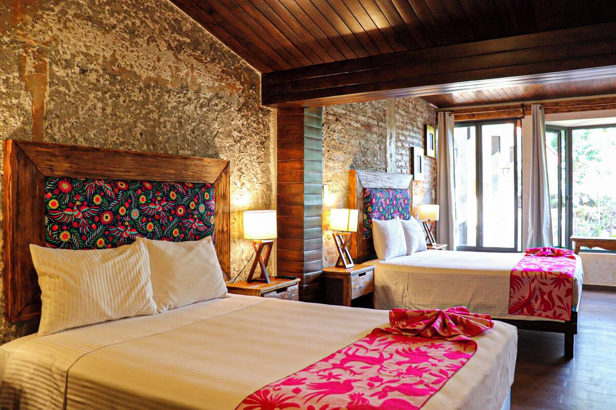 Une chambre avec deux lits, chacun doté d’une tête de lit en bois et d’une tête de lit à motifs floraux. On y trouve des tables de chevet avec des lampes et une fenêtre avec des rideaux.