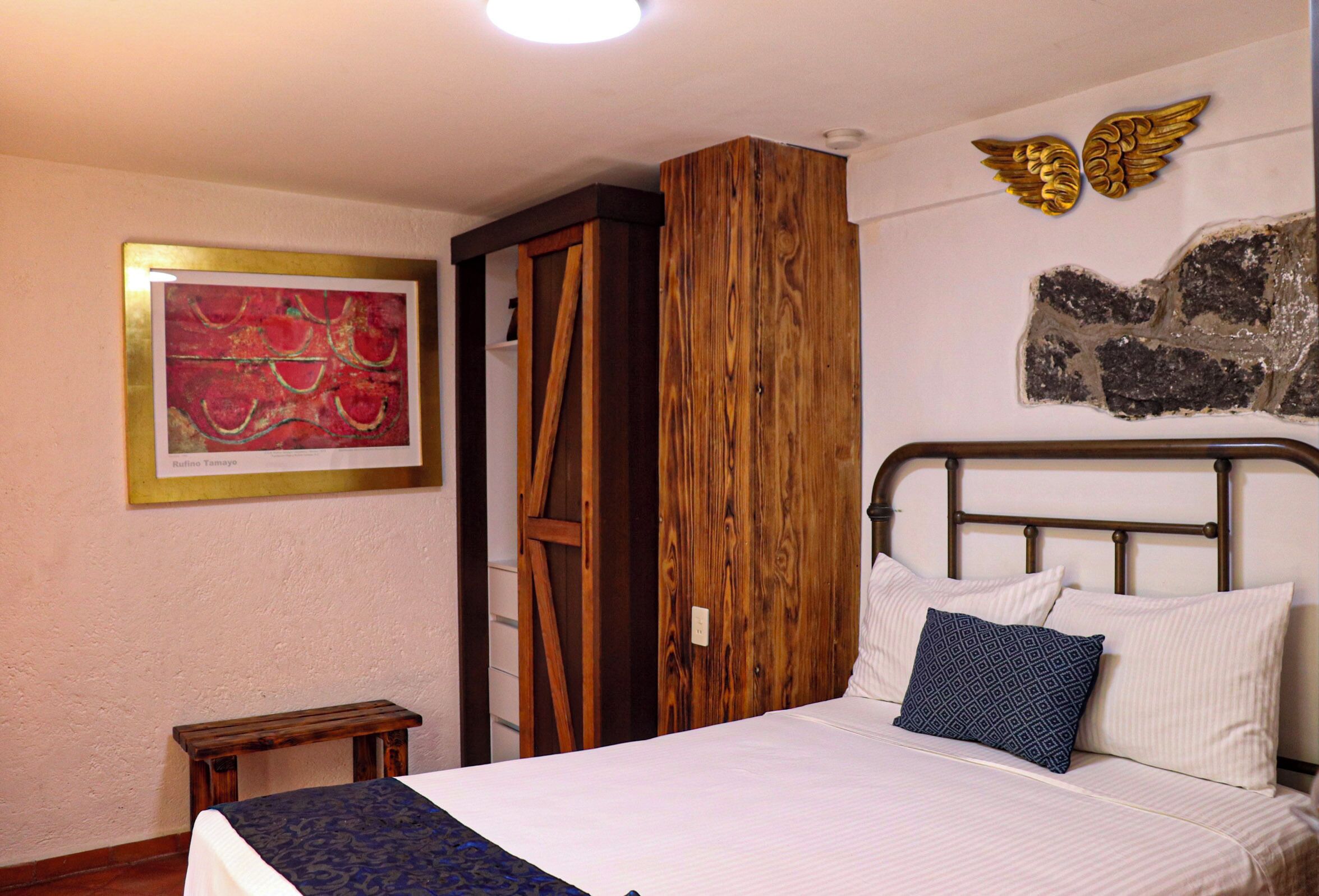 A bedroom with a bed, pillows, a small wooden stool, a wooden wardrobe, a framed artwork, and a decorative wall piece.