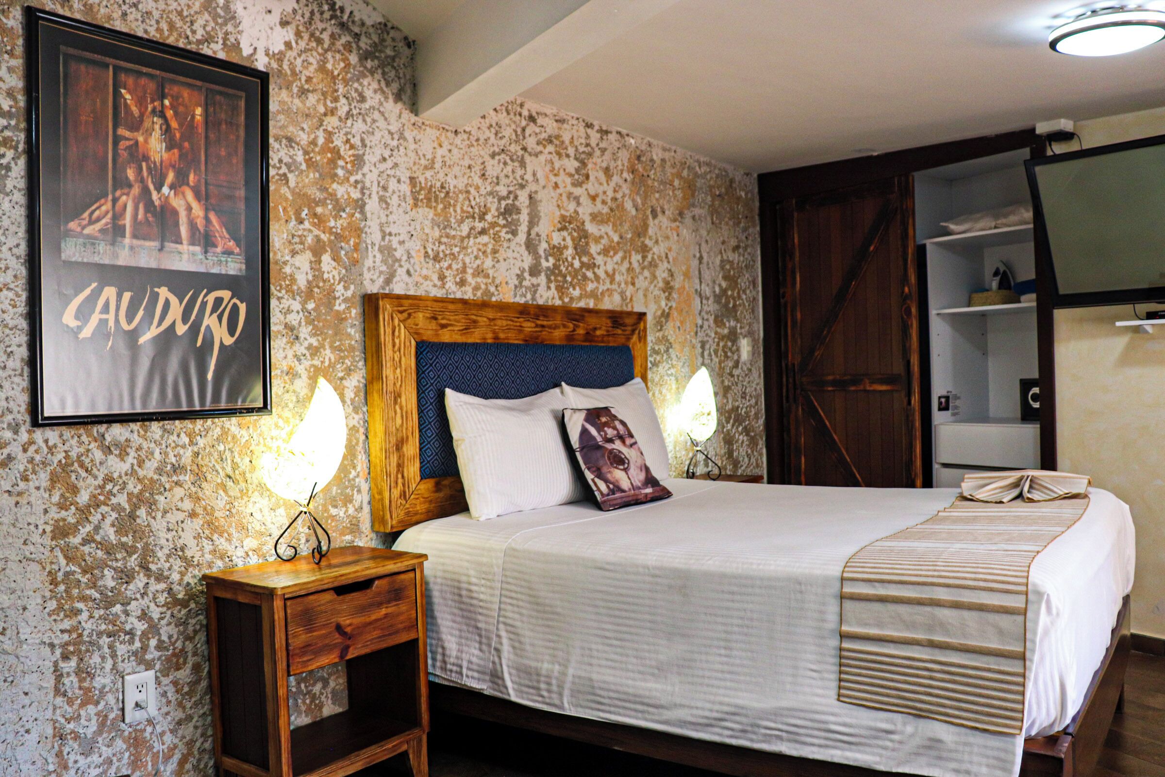 A bedroom with a wooden headboard, a bed with white bedding, a bedside table with a lamp, and a poster on the wall.