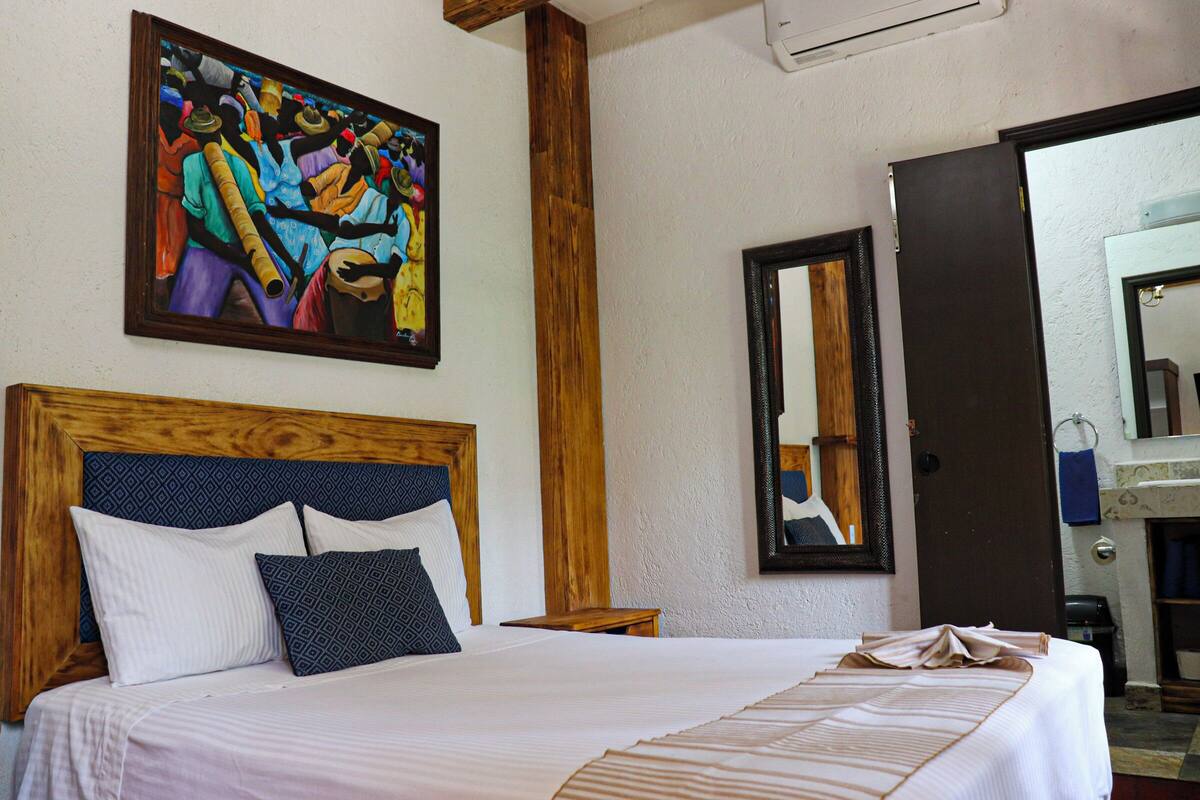 Une chambre d’hôtel avec un grand lit, une tête de lit en bois, un tableau au mur, un miroir et une porte donnant sur une salle de bain.