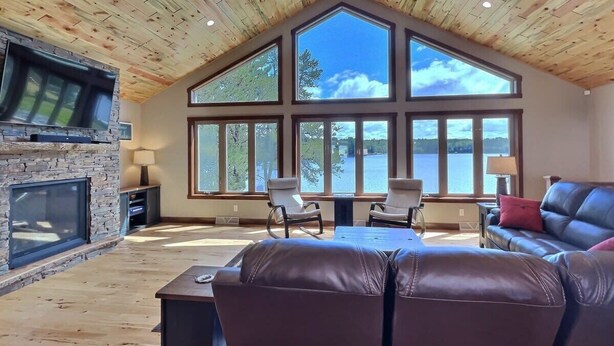 Un salón amplio con chimenea de piedra, mobiliario de cuero y ventanas grandes con vistas a un lago.