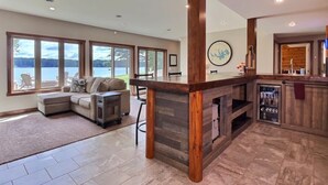 Amplia sala de estar con un gran ventanal con vista a un lago, zona de bar con encimera de madera y un espacio de descanso con sofá y sillones.