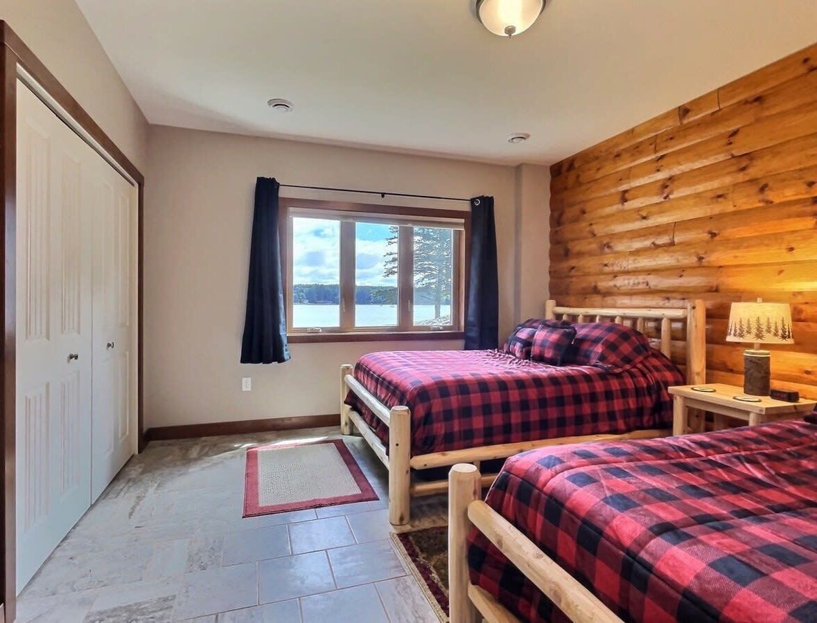 A bedroom with two beds, a window with curtains, a wooden wall, and a nightstand with a lamp.